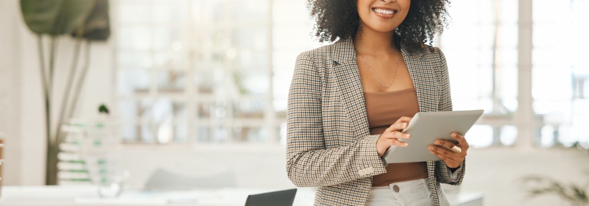 Smiling woman in a blazer holding a tablet, sitting at a desk with a laptop in a bright office.